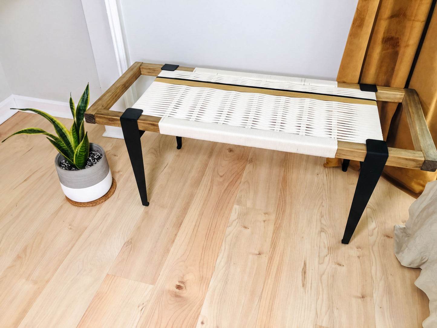 a wooden bench with a woven seat and black metal legs, placed on a wooden floor next to a potted plant.
