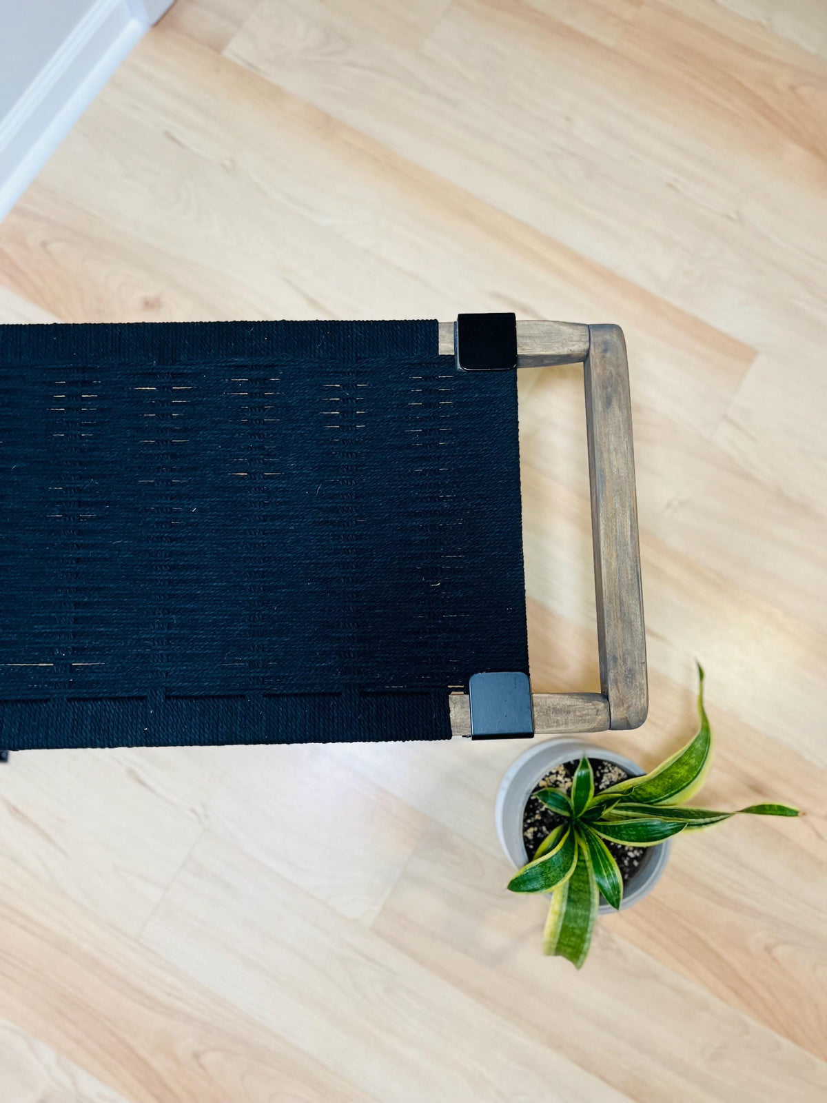 a black woven bench with a wooden handle, placed on a wooden floor next to a small potted plant.