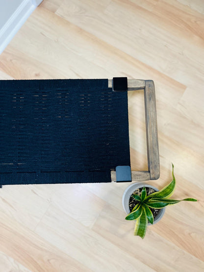 a black woven bench with a wooden handle, placed on a wooden floor next to a small potted plant.