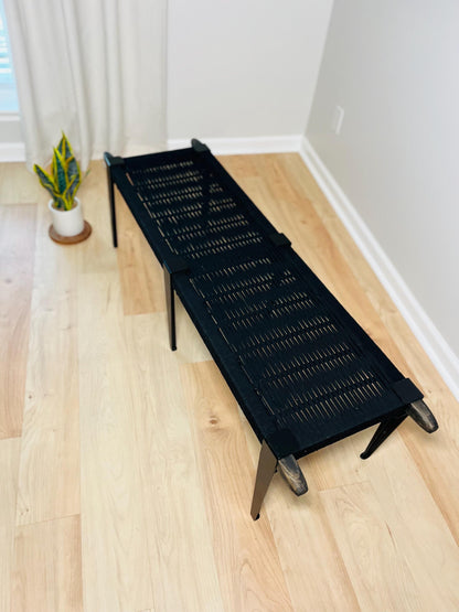 a black woven bench with a wooden frame, placed on a wooden floor next to a window with white curtains.