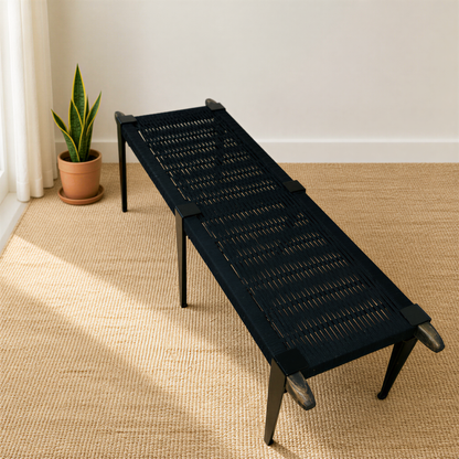 a black woven bench with a woven seat and legs, placed on a beige carpeted floor next to a potted plant.