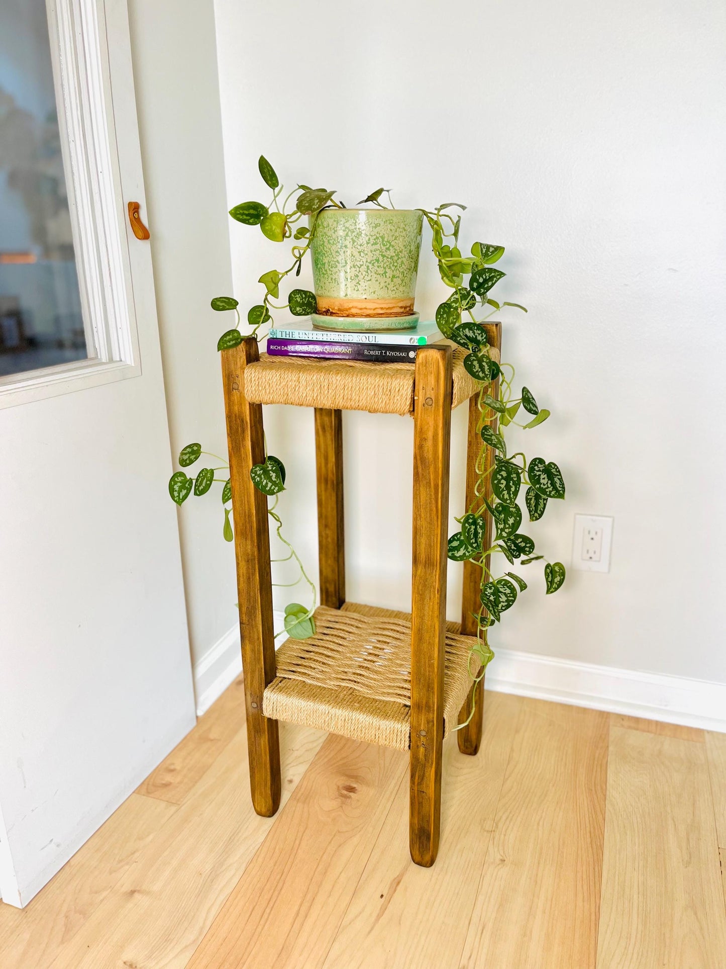 A wooden side table with a woven seat and a plant on top, situated in a corner of a room with a white wall and a window.