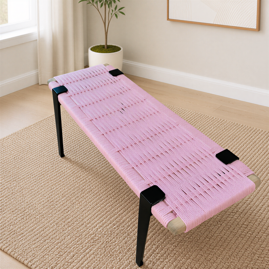 A pink woven bench with black metal legs sits on a beige rug in a room with a window and a plant.
