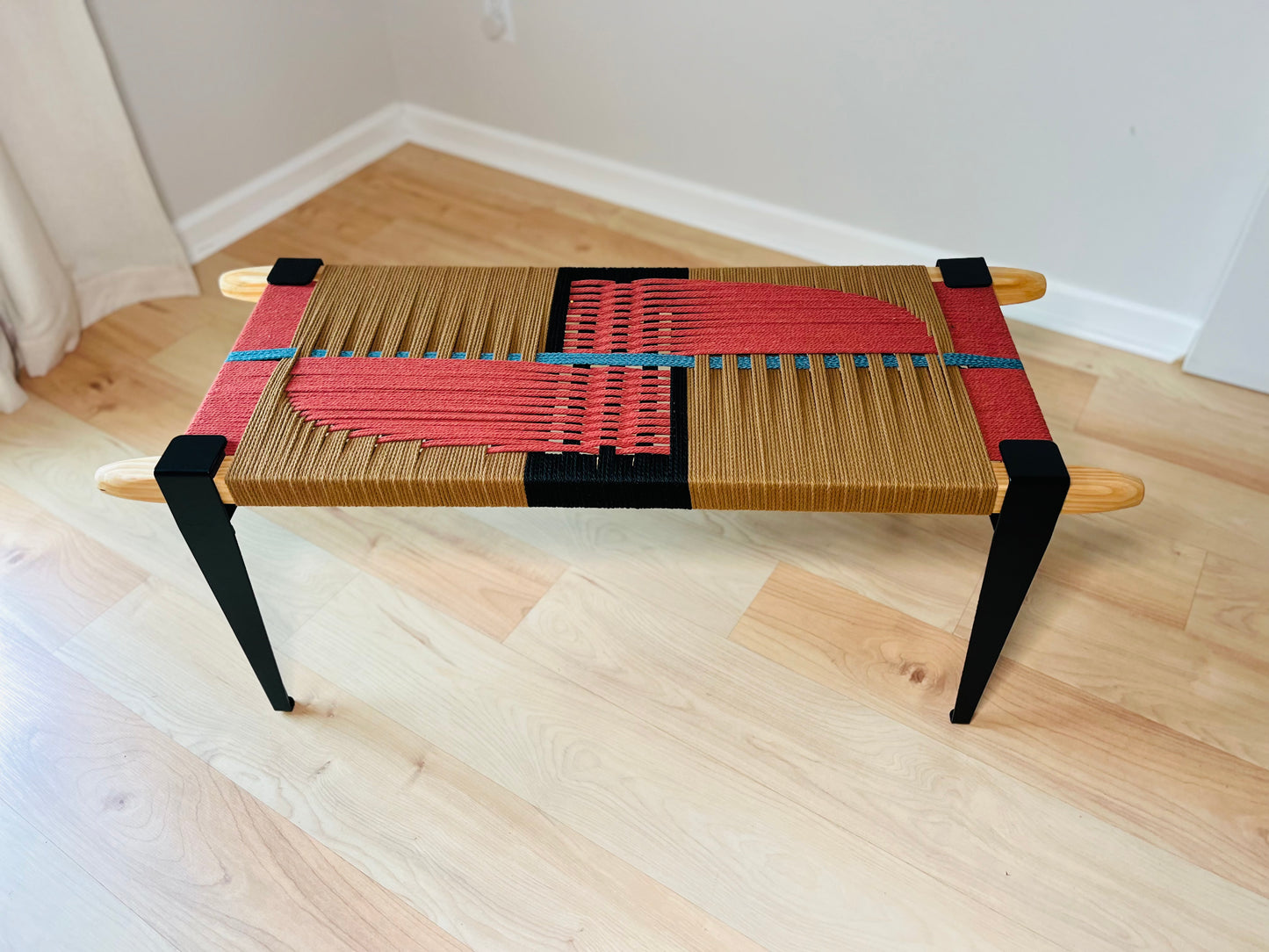 a modern, wooden bench with a woven seat and legs. The bench has a striking design with red and black woven elements, creating a visually appealing contrast. The bench is placed on a wooden floor, which complements the overall aesthetic of the piece.