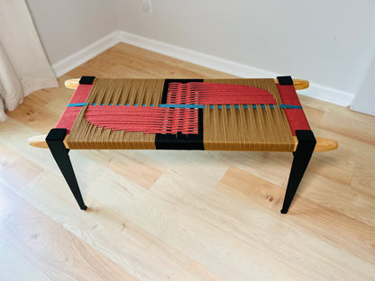 a modern, wooden bench with a woven seat and legs. The bench has a striking design with red and black woven elements, creating a visually appealing contrast. The bench is placed on a wooden floor, which complements the overall aesthetic of the piece.