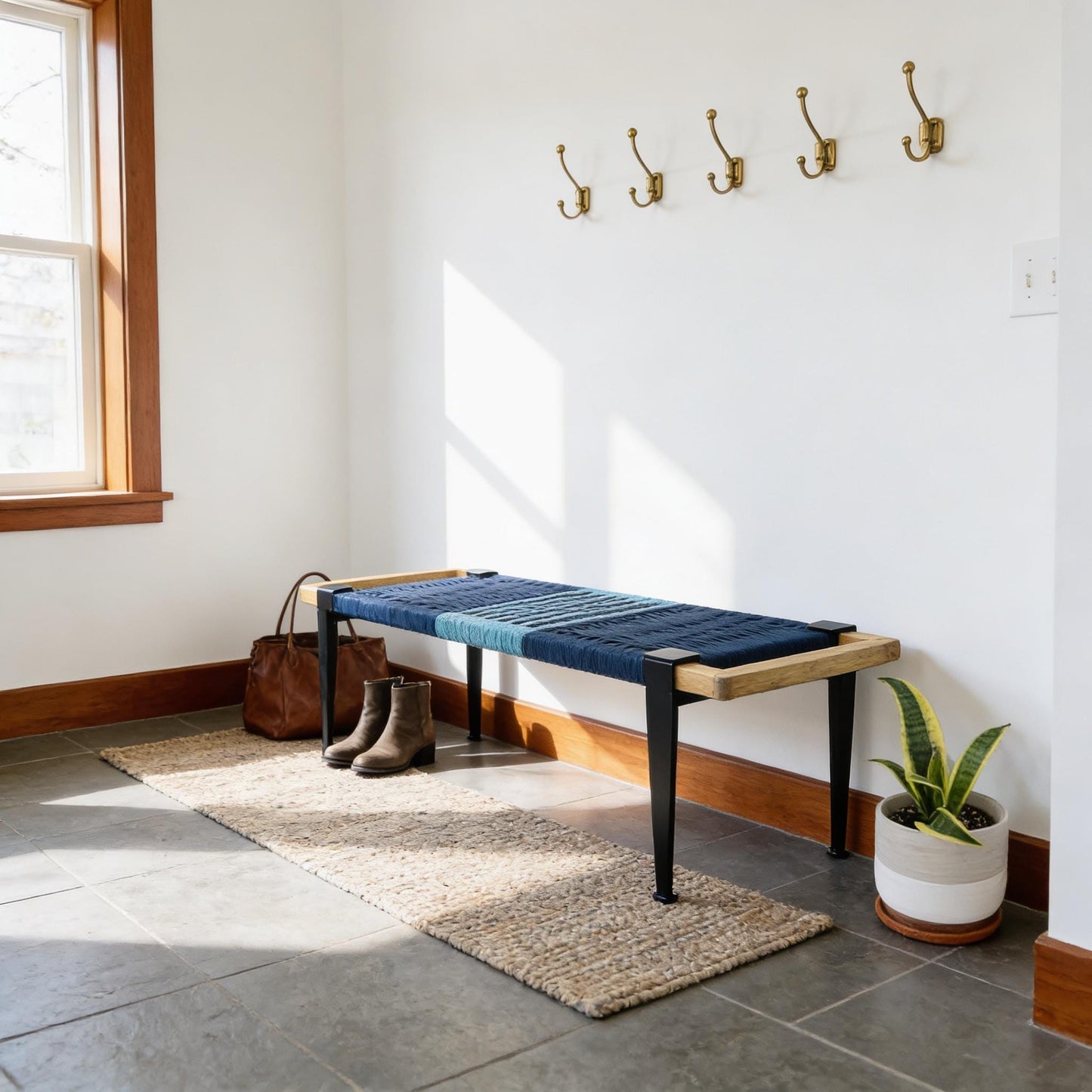 a hallway with a blue bench, a brown leather bag, and a potted plant. The bench is positioned against a white wall with a window, and there are four gold coat hooks mounted on the wall above the bench.
