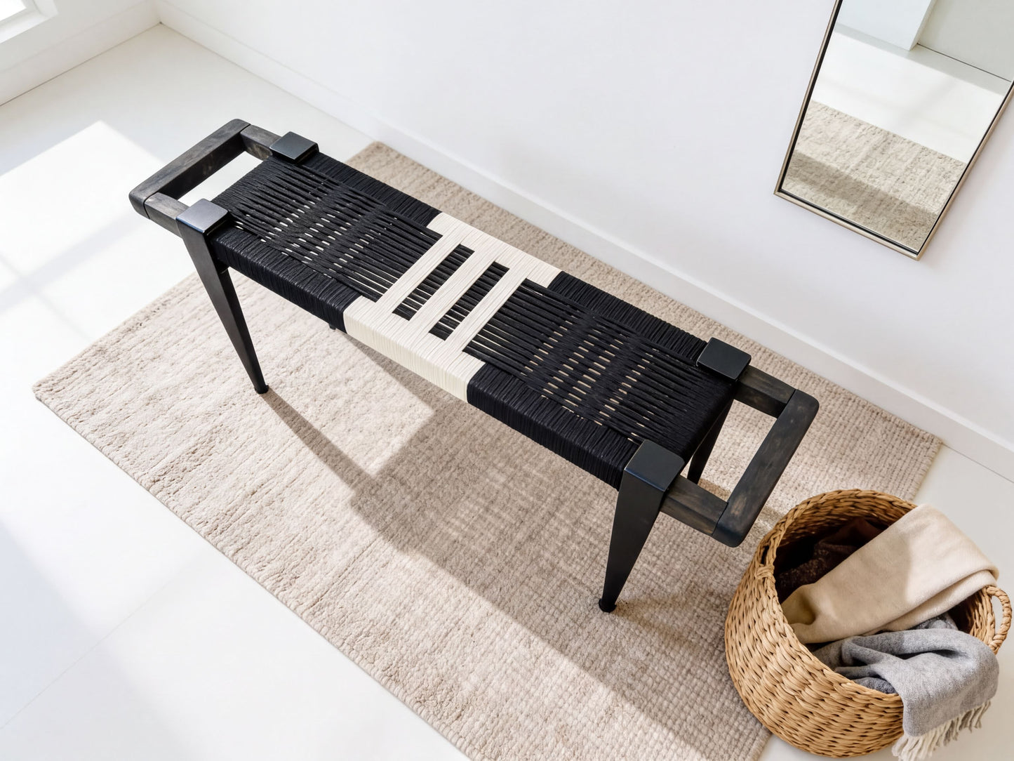 a black and white bench with a woven seat and black metal legs, placed on a beige rug. Next to the bench is a woven basket filled with various items, including a gray blanket and a beige scarf.