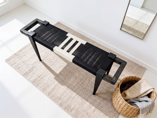 a black and white bench with a woven seat and black metal legs, placed on a beige rug. Next to the bench is a woven basket filled with various items, including a gray blanket and a beige scarf.