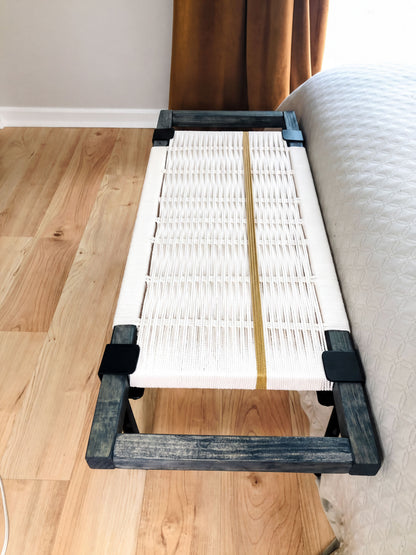 a white woven bench with a wooden frame, placed on a wooden floor next to a bed.