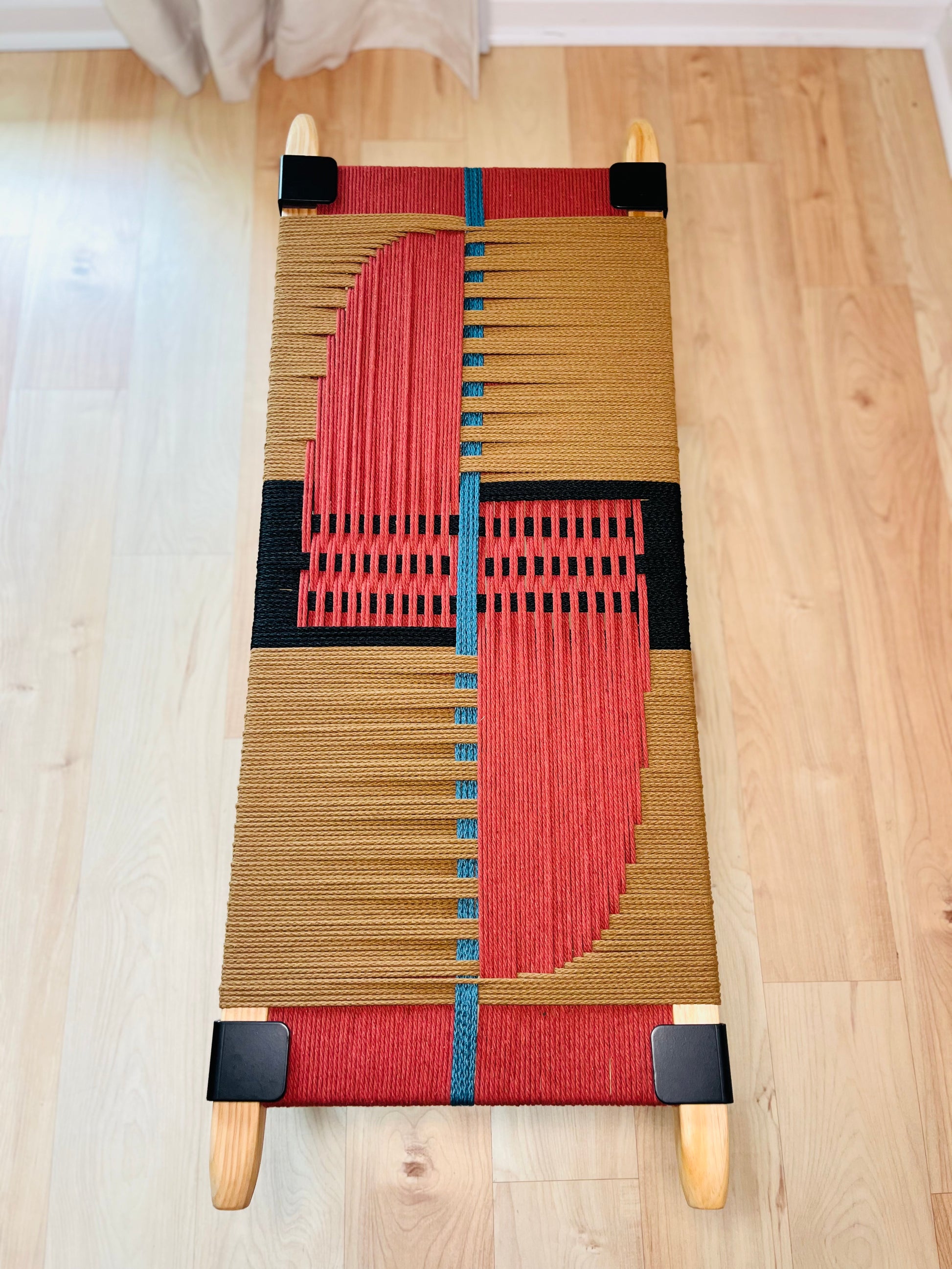 a woven bench with a striking geometric design in red, black, and blue colors. The bench is placed on a wooden floor, and there are no other objects or people visible in the image.