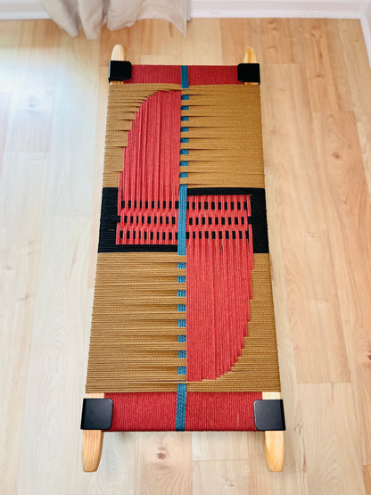 a woven bench with a striking geometric design in red, black, and blue colors. The bench is placed on a wooden floor, and there are no other objects or people visible in the image.