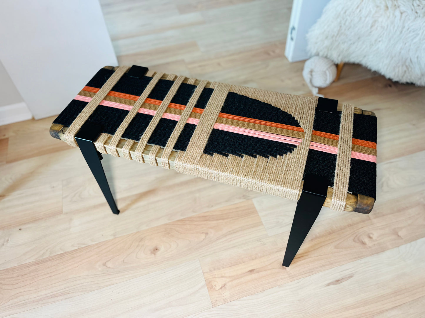 A bench with a woven seat and legs, featuring a geometric pattern in black, orange, and pink colors. The bench is placed on a wooden floor, and there is a white furry object in the background.