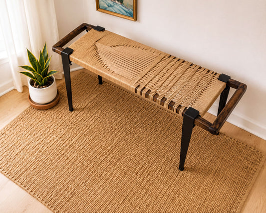 a woven bench with a woven seat and wooden frame, placed on a woven rug in a room with a window and a painting on the wall.