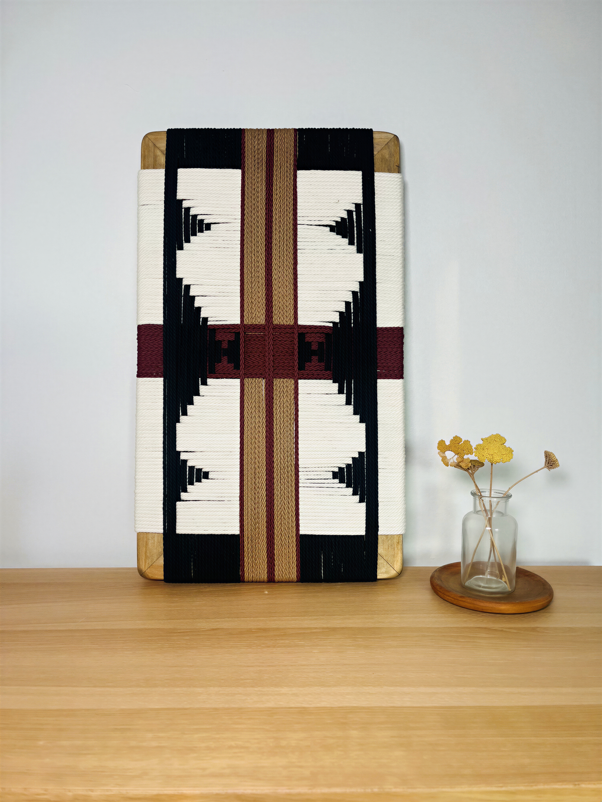 a wooden table with a decorative woven piece, a vase with yellow flowers, and a wooden coaster.