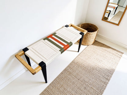 a modern bench with a wooden frame and a woven seat, placed on a beige rug in a corner of a room with a mirror on the wall.