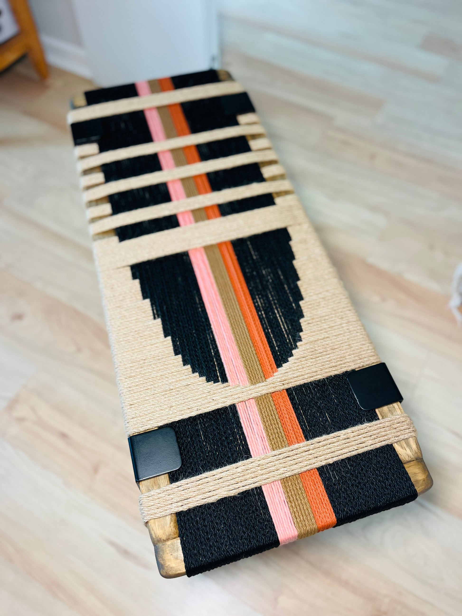 a wooden bench with a unique, colorful design made of rope and fabric. The bench has a black and white striped pattern, with orange and pink stripes running through it. The bench is placed on a wooden floor, and there is a wooden shelf visible in the background.