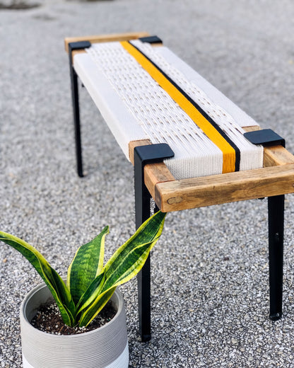 a wooden bench with a woven seat and metal legs, placed on a gravel surface next to a potted plant.