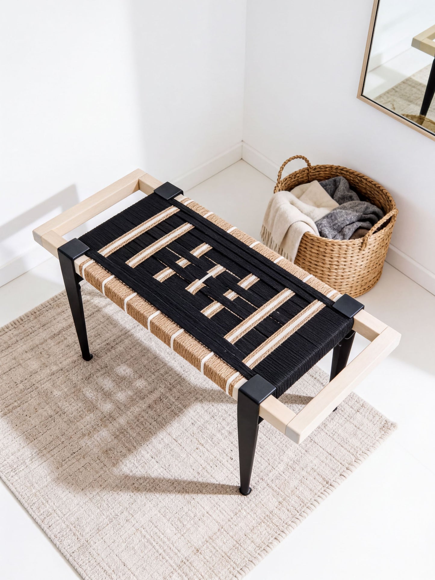 a black and white bench with a woven seat and black metal legs, placed on a beige rug in a room with a basket of clothes nearby.