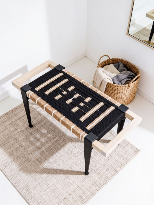 a black and white bench with a woven seat and black metal legs, placed on a beige rug in a room with a basket of clothes nearby.
