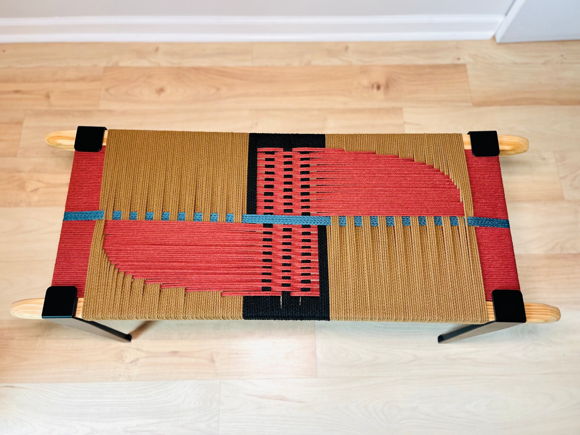 a woven bench with a geometric pattern in red, brown, and black colors. The bench is placed on a wooden floor, and it appears to be a modern and stylish piece of furniture.