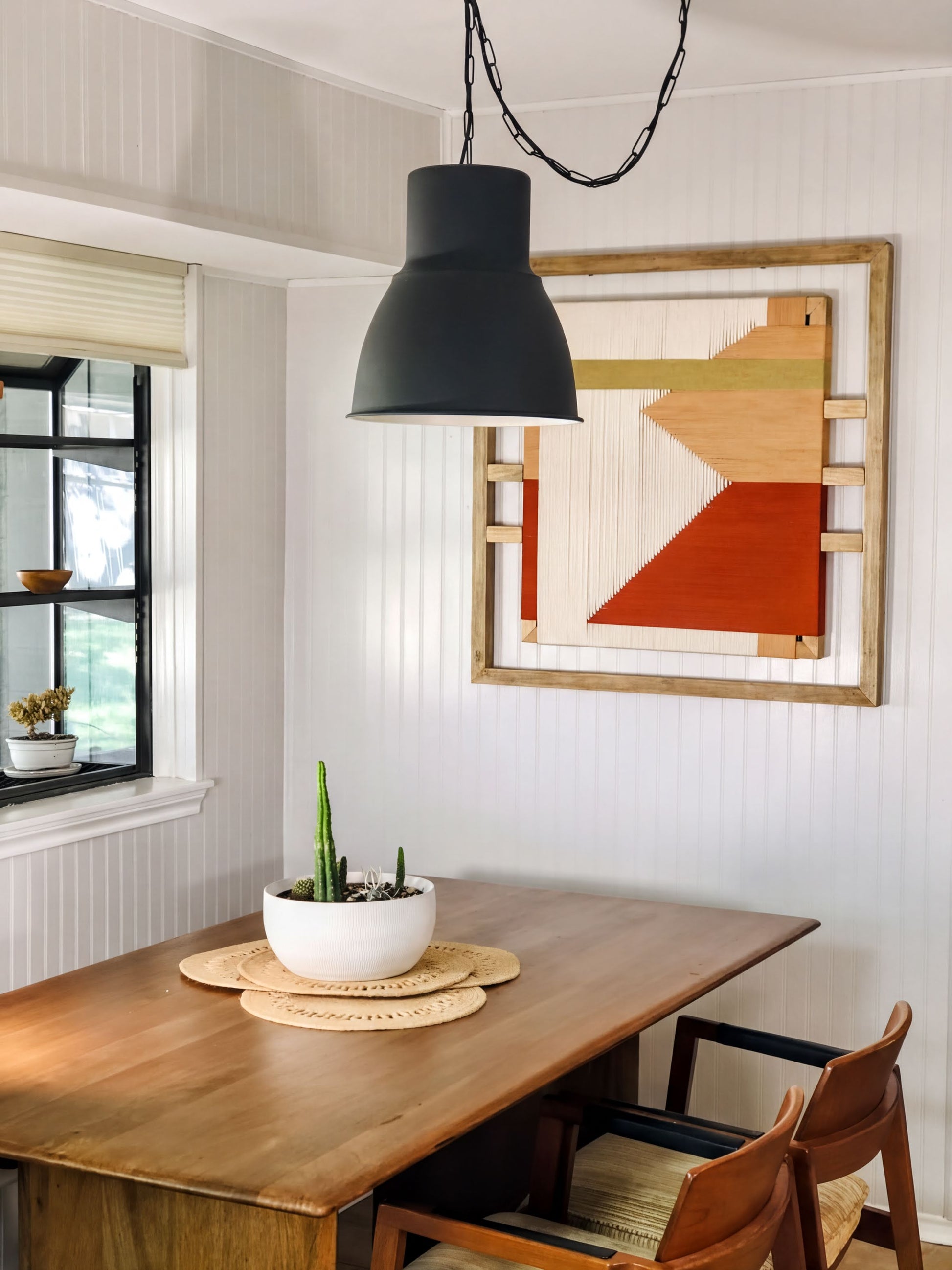 a dining room with a wooden table, a potted plant, and a hanging light fixture. The table is set with a bowl and a plant, creating a cozy and inviting atmosphere.