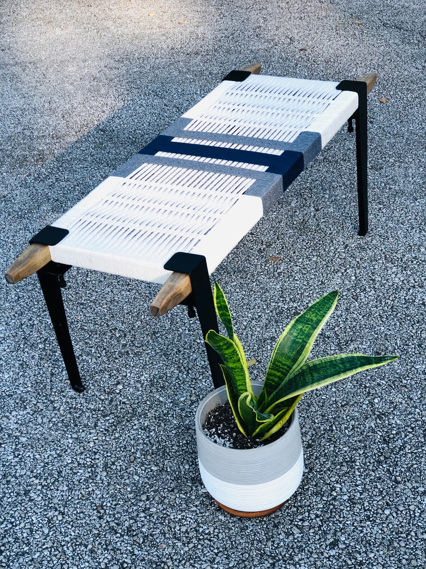 a white bench with a black metal frame, placed on a gravel surface next to a potted plant.
