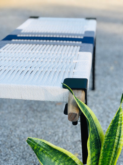 a white woven bench with a wooden support, placed on a concrete surface. A potted plant is visible in the foreground, adding a touch of greenery to the scene.